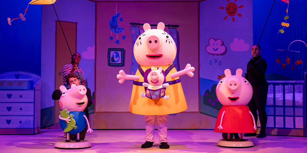 George, Mummy Pig, Evie, and Peppa Pig stand together, smiling, in a colourful stage setting.