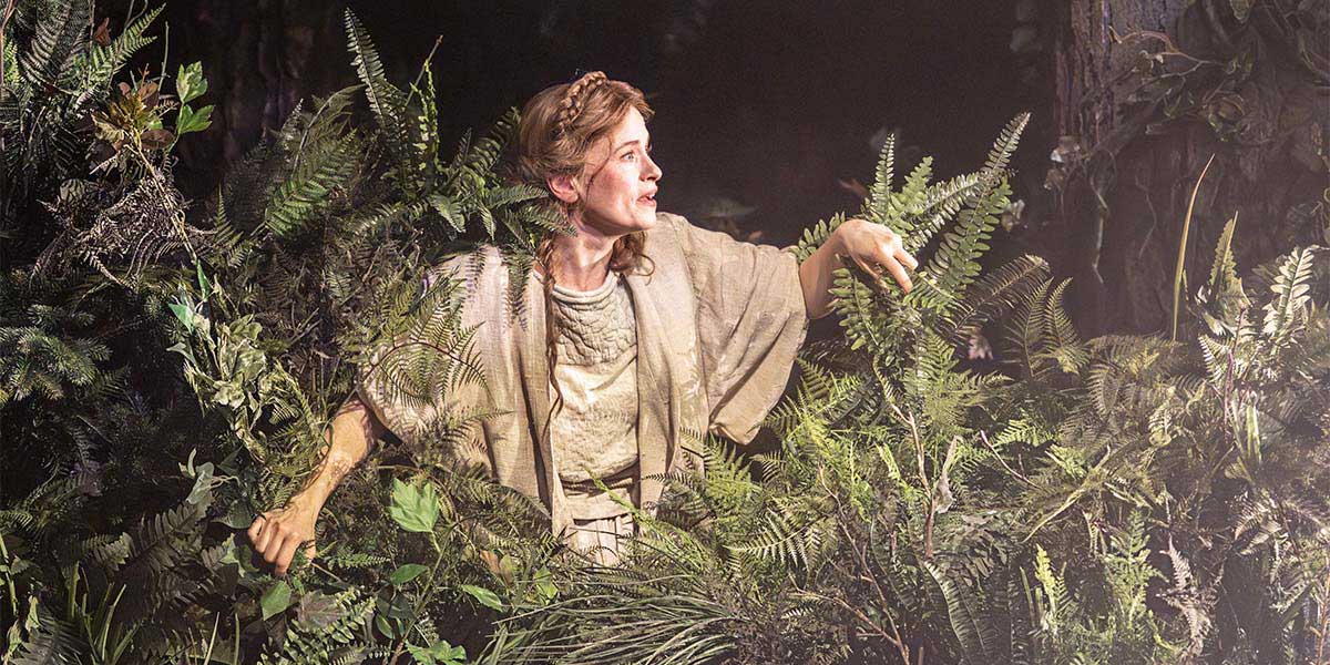 Katie Brayben in a forest setting, wearing a green dress, surrounded by trees and soft lighting.