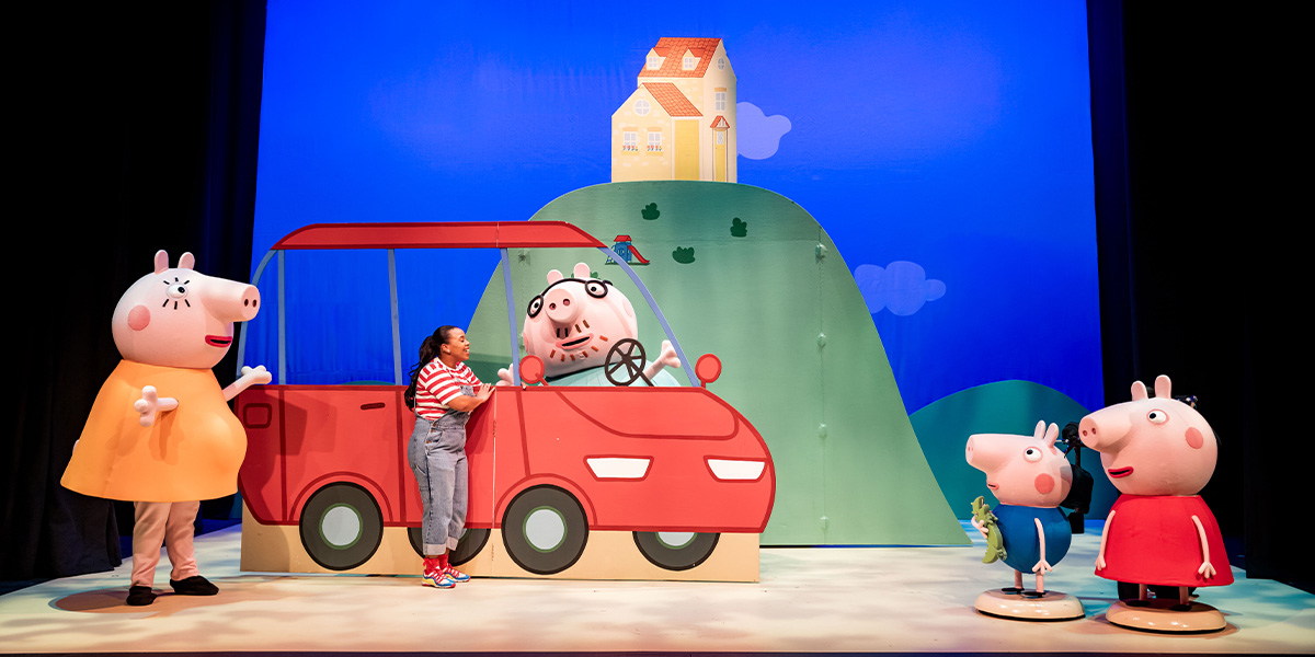 Mummy, Daisy, Daddy, George, and Peppa Pig are smiling beside a colourful car at a show.