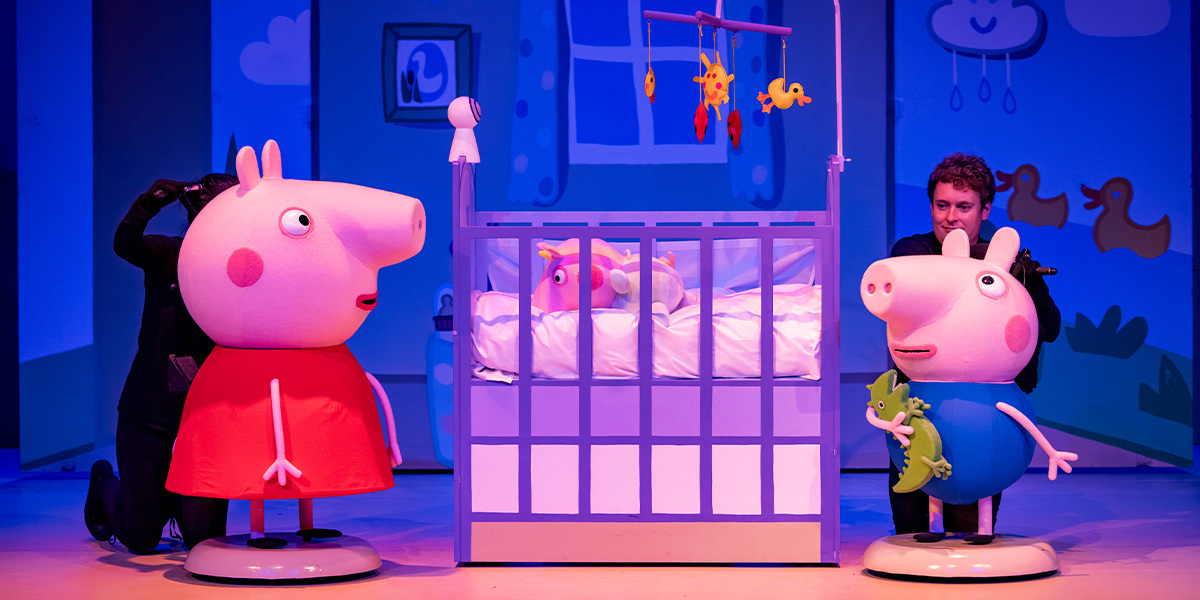 Peppa Pig, Evie, and George are playing together in a colourful crib, surrounded by soft toys.