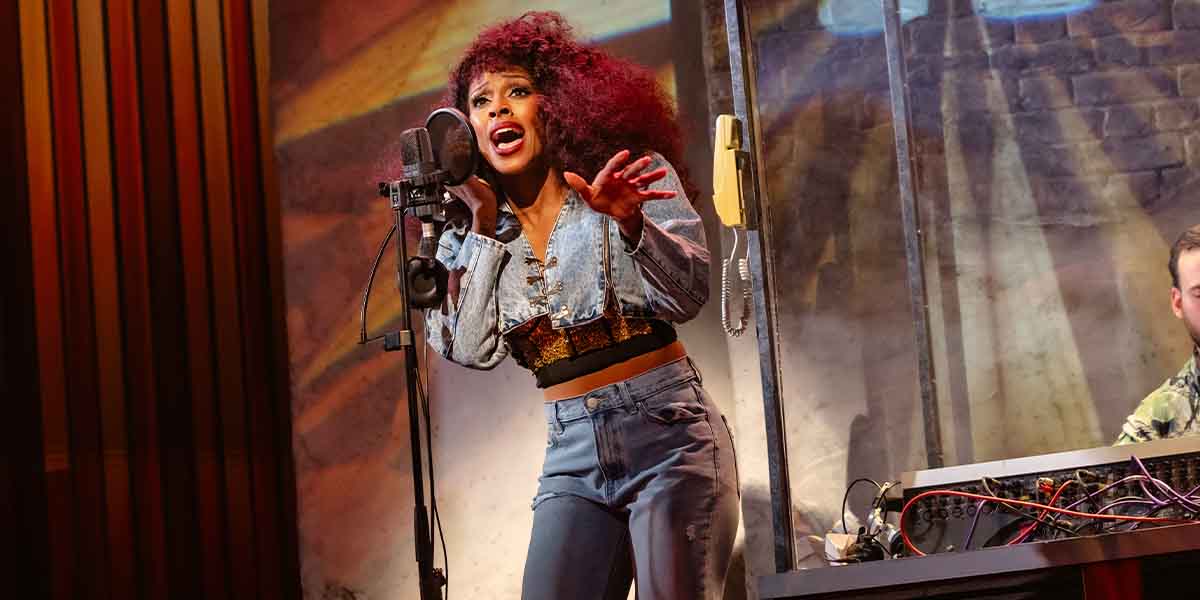 A person with curly hair sings passionately into a microphone, wearing a denim jacket and jeans.