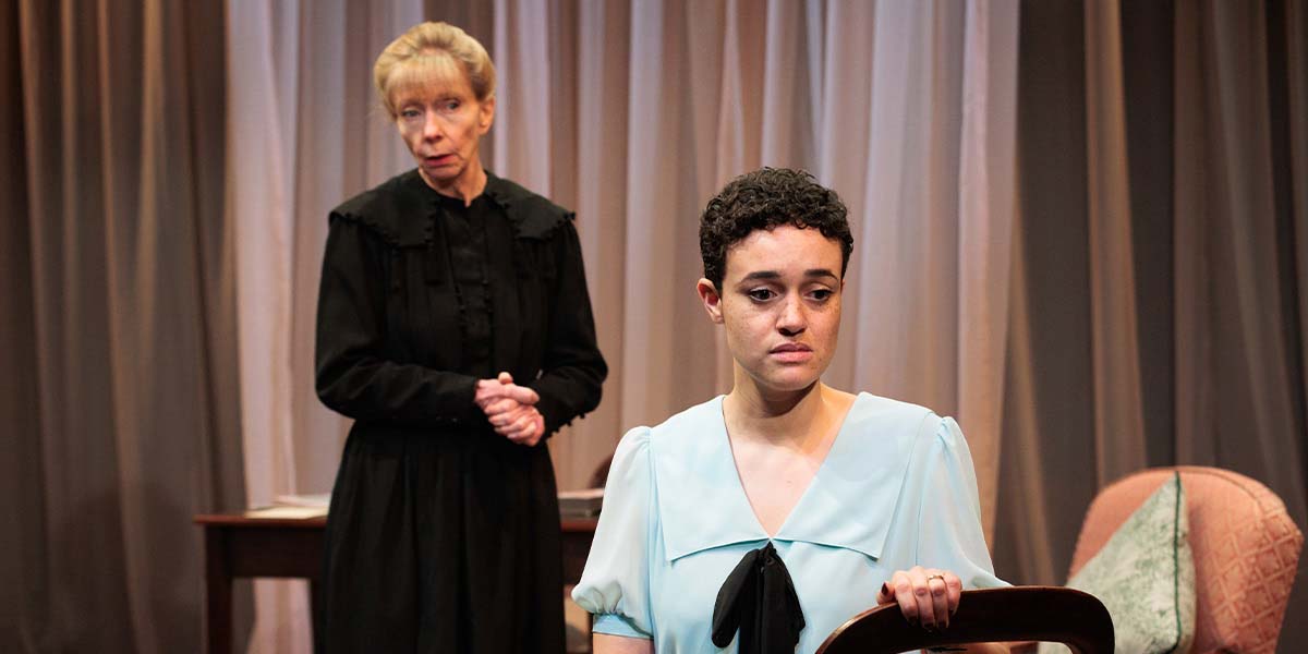 Angela Sims stands in black attire, while Lily Nichol, in a light blue dress, sits, looking distressed.