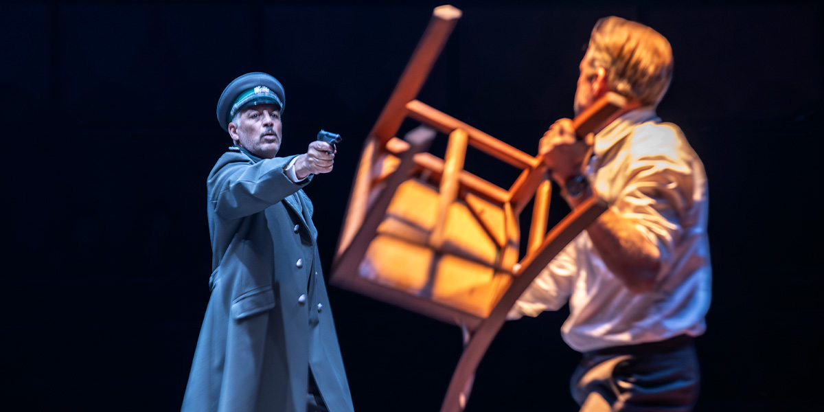 David Rubin points a gun at Rory Keenan, who raises a wooden chair, in a dramatic scene.