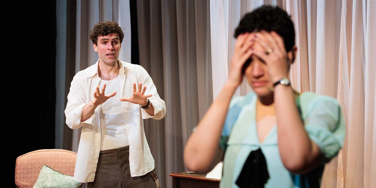 Ewan Miller gestures expressively while Lily Nichol covers her face, conveying tension in a theatrical scene.