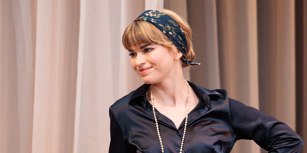 Gina Bramhill smiles while wearing a dark blouse and floral headscarf, with draped curtains in the background.