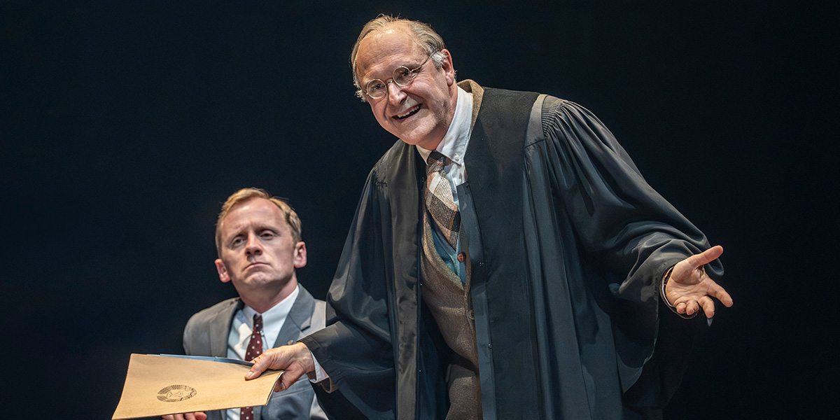 Gunnar Cauthery smiles while gesturing with a folder, as John Ramm looks on, serious.