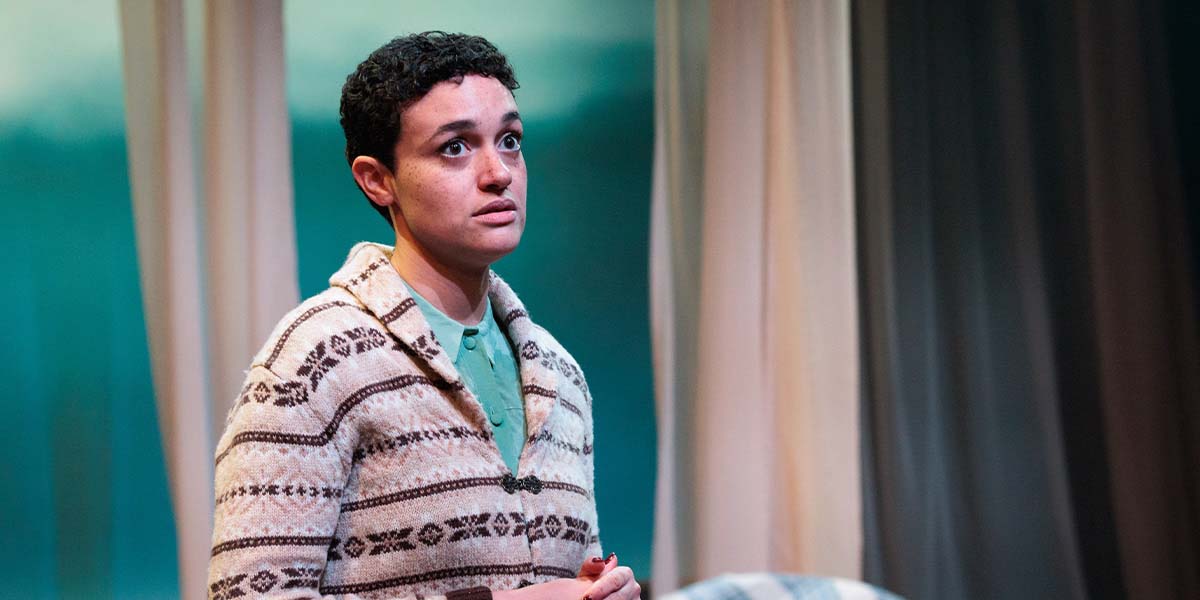 Lily Nichol, wearing a patterned cardigan, looks concerned while standing in a softly lit stage setting.