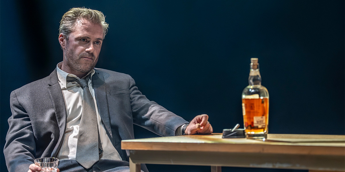 Rory Keenan portrays a character in a dramatic pose, dressed in a dark coat against a muted background.