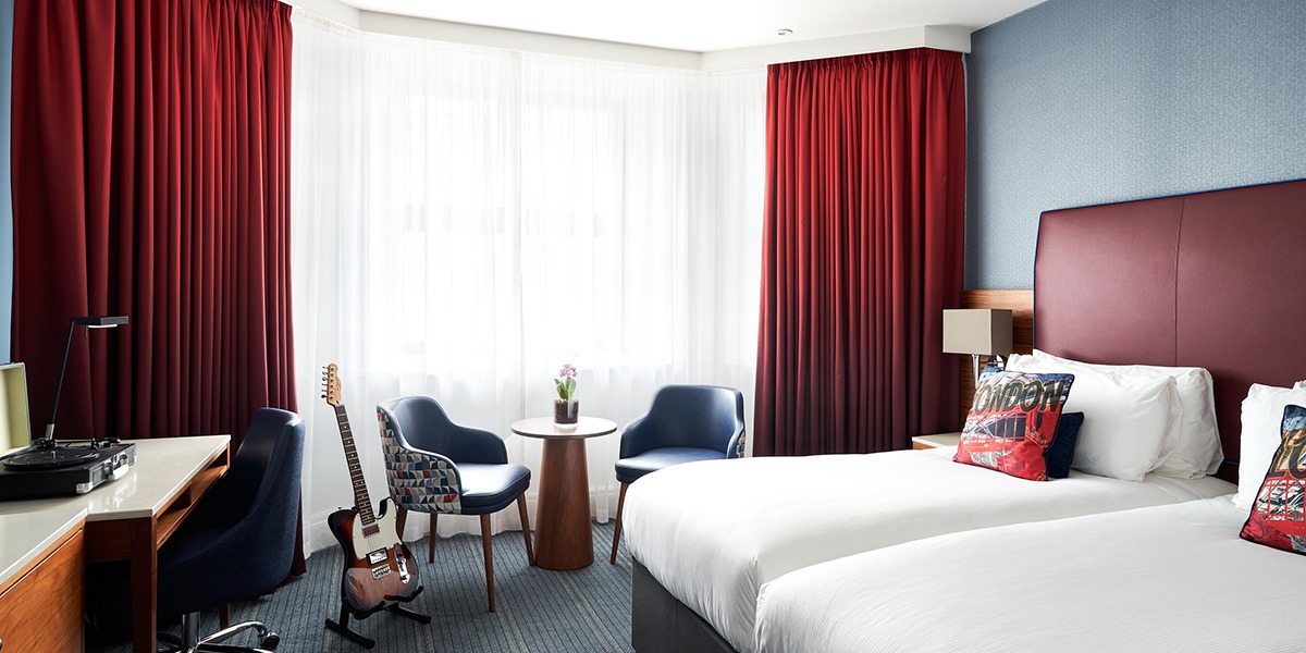 A hotel bedroom with twin single beds and sidetable, plus an electric guitar in a stand and a music turntable on a desk