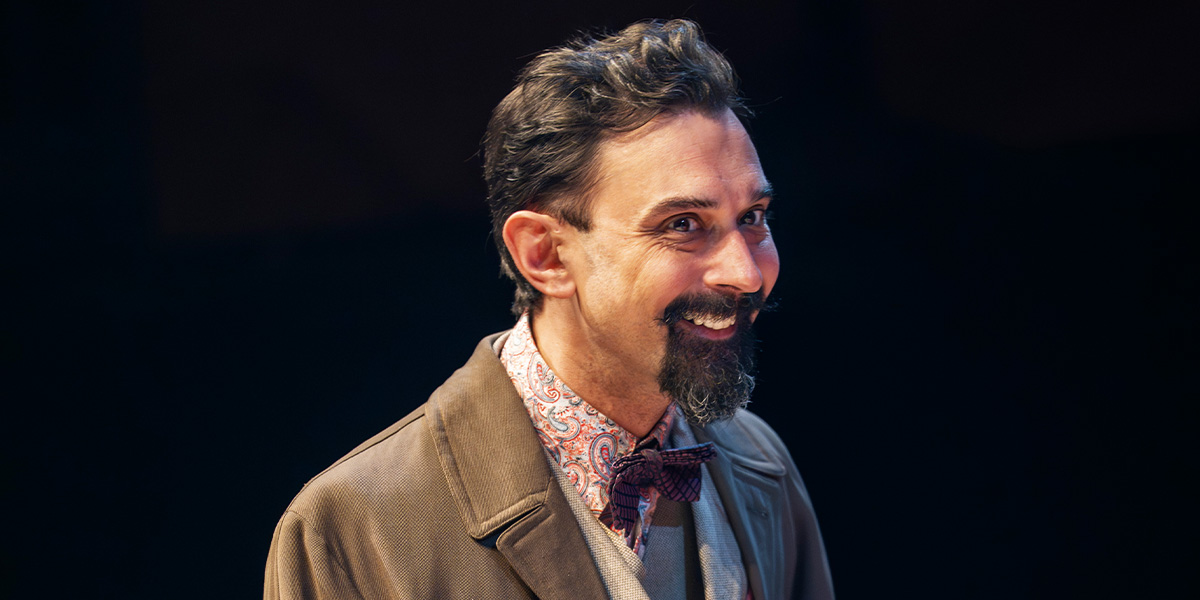 Tom Kanji smiles while wearing a patterned shirt and bow tie, dressed in a brown coat.