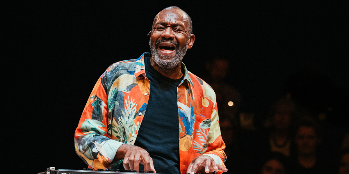 Lenny Henry sits on a stool, smiling, wearing a black shirt, with a colourful backdrop behind him.