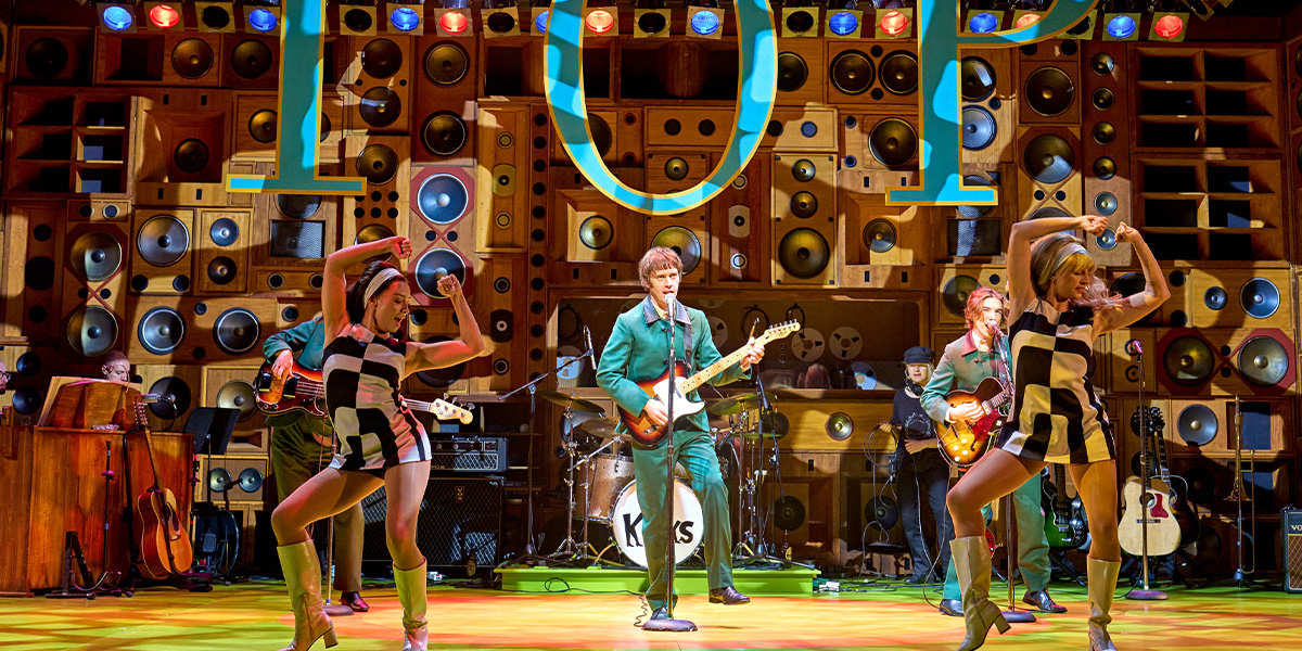 A group of performers in vibrant costumes are singing energetically on stage, with bright lighting and a cheering audience.