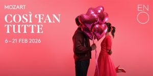Two elegantly dressed women in 18th-century attire interact on a stage, with ornate curtains in the background.