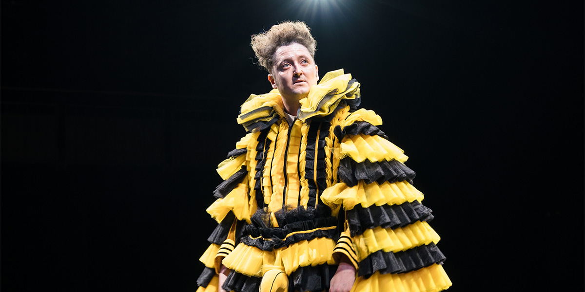A performer in a dramatic yellow and black costume stands under a spotlight, gazing intently.