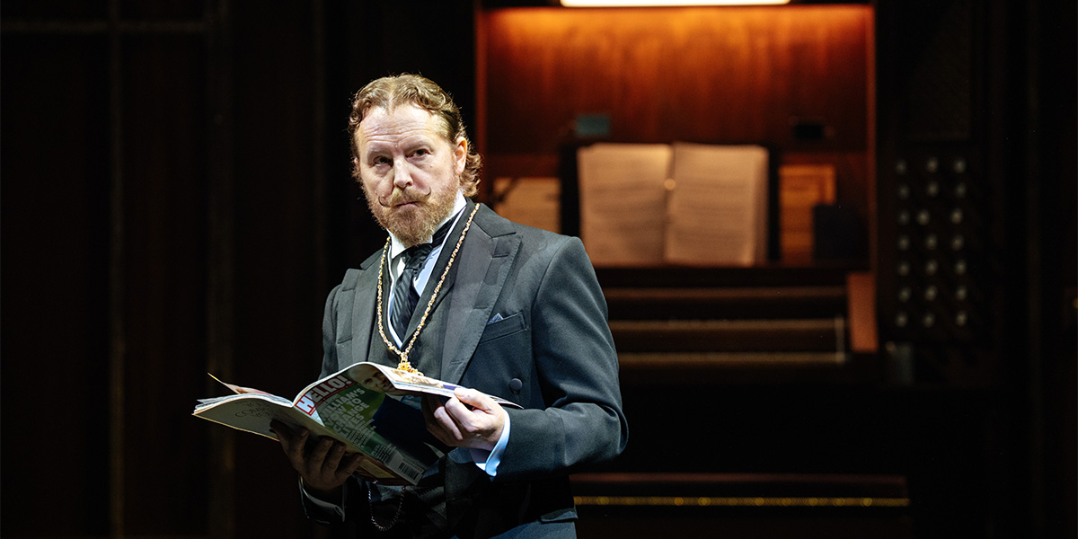 A person in formal attire reads a magazine, standing in front of a wooden backdrop with music sheets.
