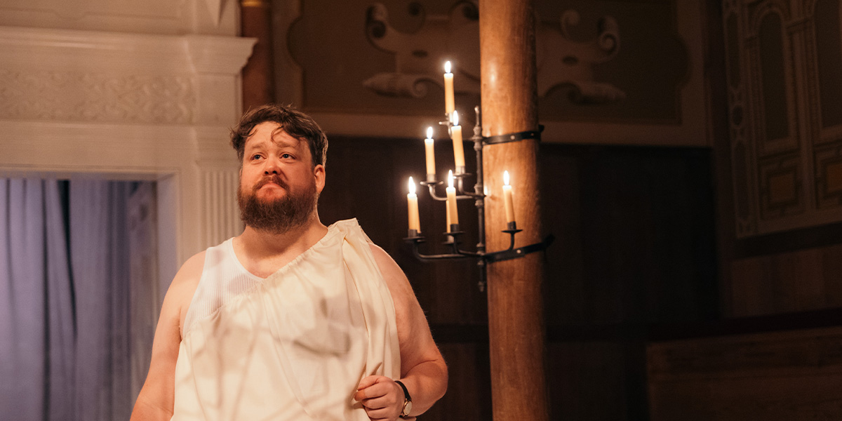 A man in a white tunic stands under candlelight, looking thoughtfully upwards in a theatrical setting.