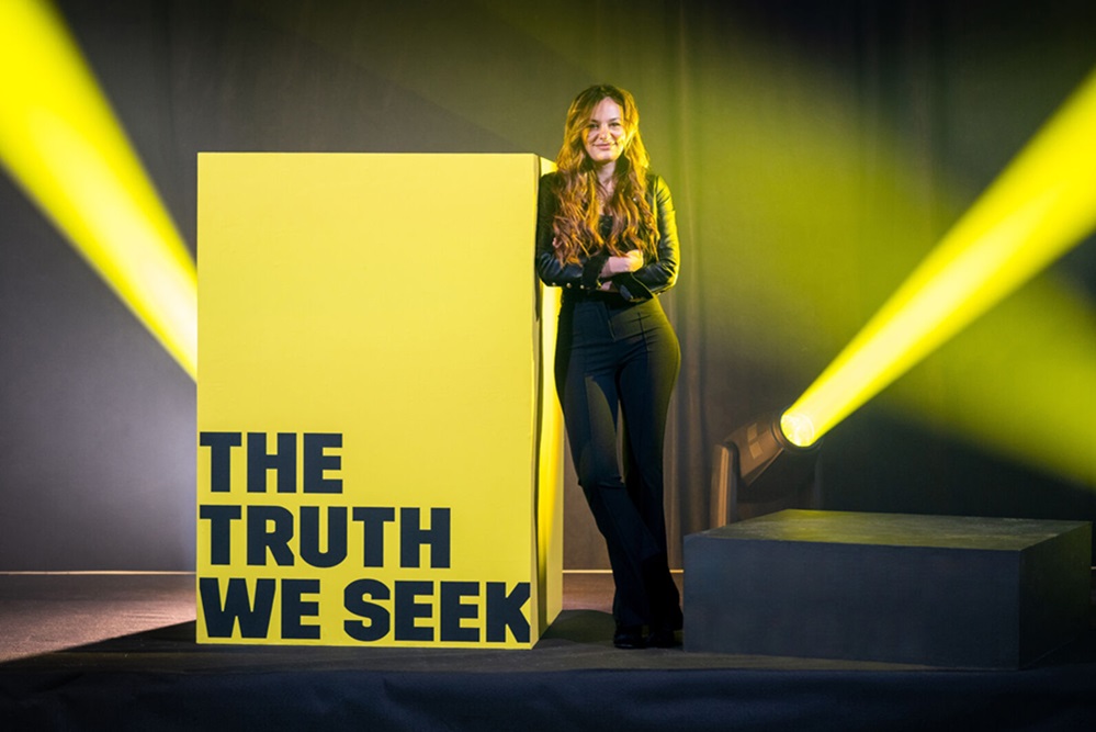 A woman standing next to a yellow box that reads: The Truth We Seek