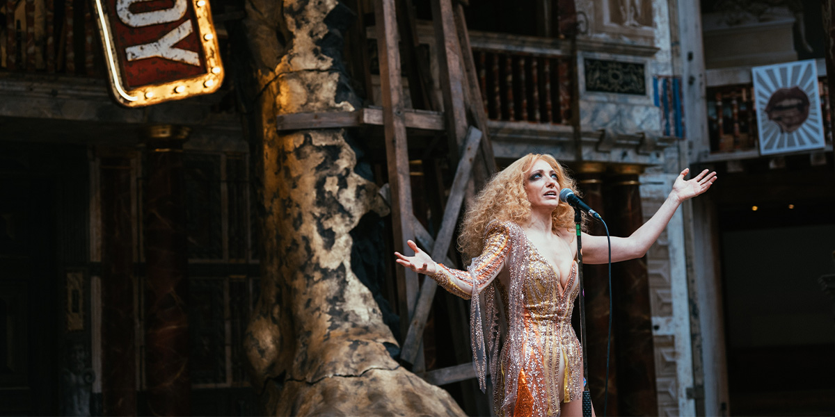 Performer singing on stage at the Globe Theatre, surrounded by theatrical props and a sign with the word "Troy."
