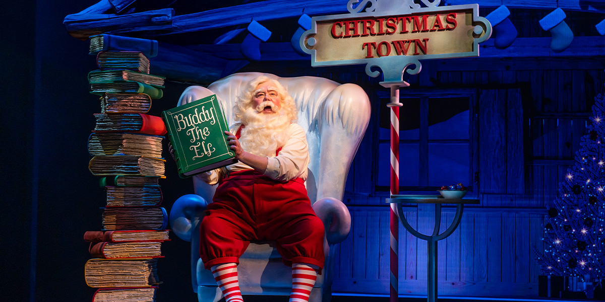 Martyn Ellis dressed as Santa Claus, smiling, surrounded by festive decorations in a theatrical setting.
