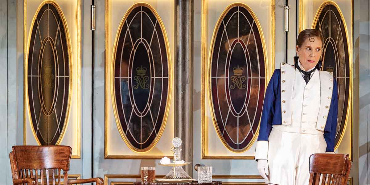 Mel Giedroyc stands in a white and blue costume, with ornate glass panels in the background.