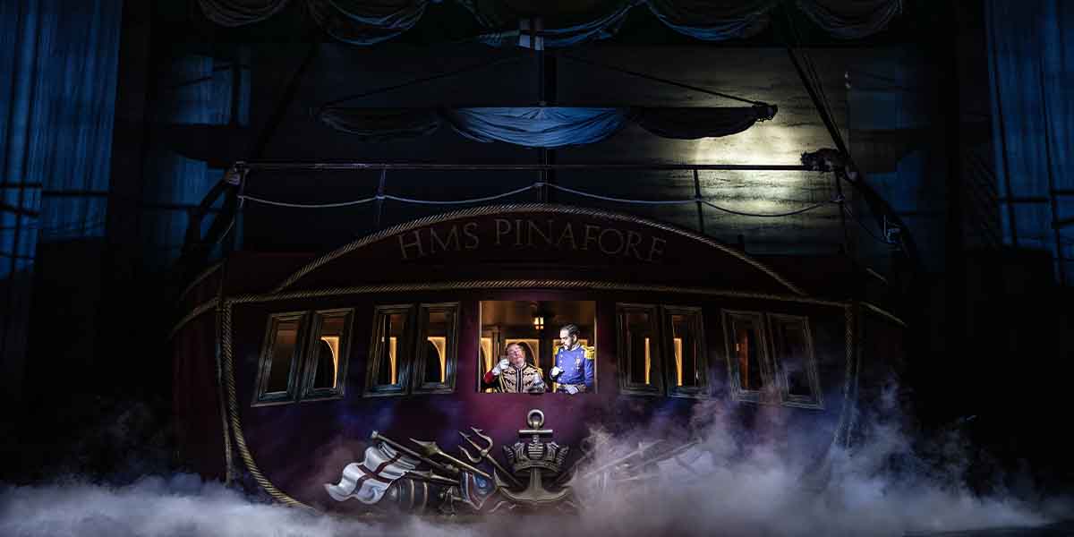 Neal Davies and John Savournin perform on stage in front of the HMS Pinafore set, shrouded in fog.