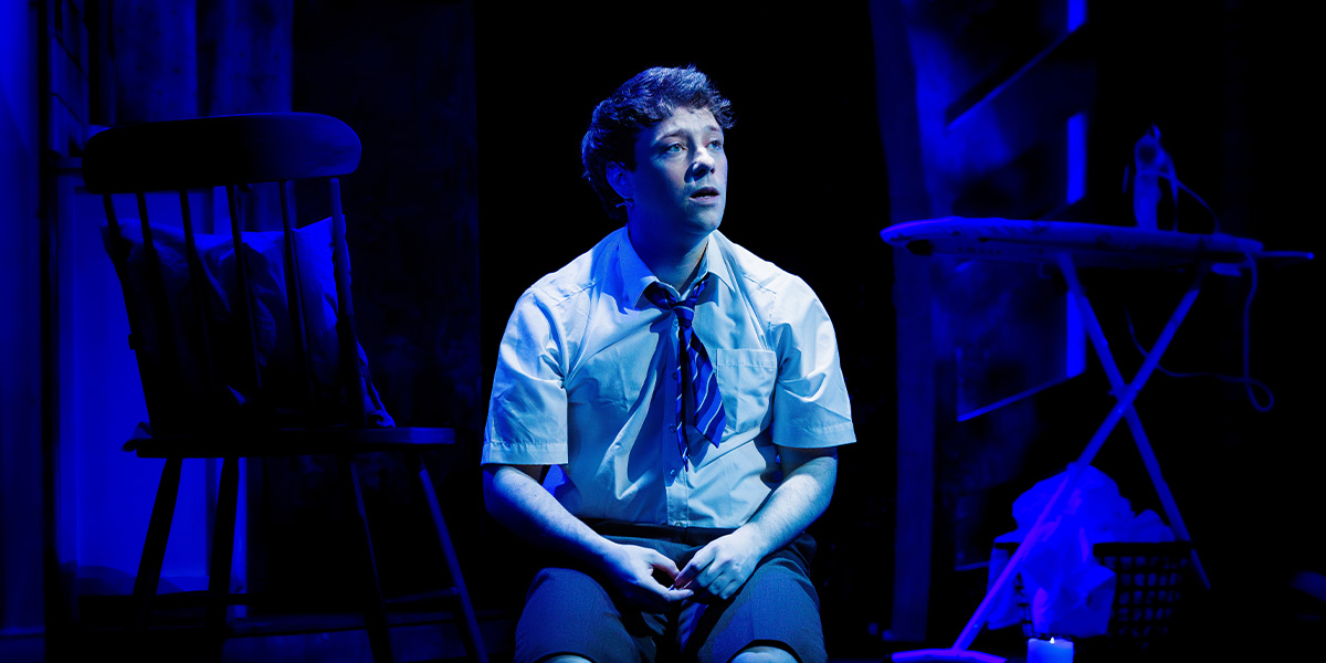Andy Owens sits on stage, looking contemplative, wearing a white shirt and blue tie, with blue lighting.