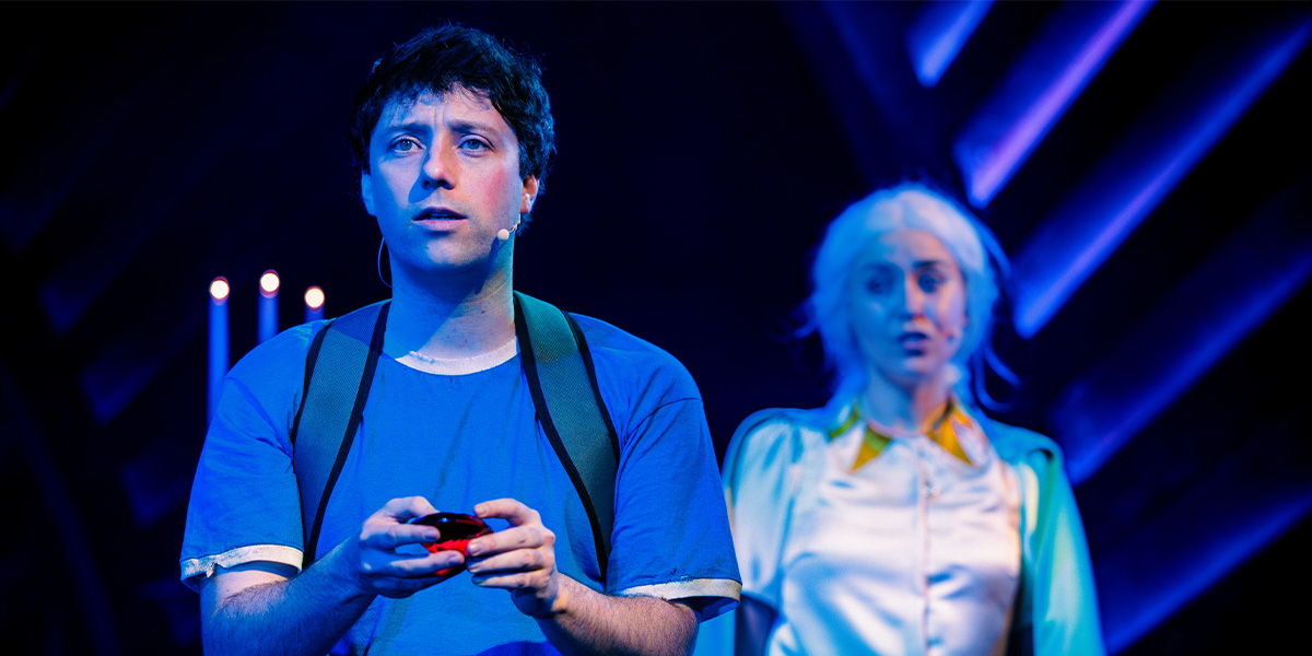 Andy Owens holds a gaming controller, looking concerned, while Nadia Wyn Abouayen stands behind him, surprised.