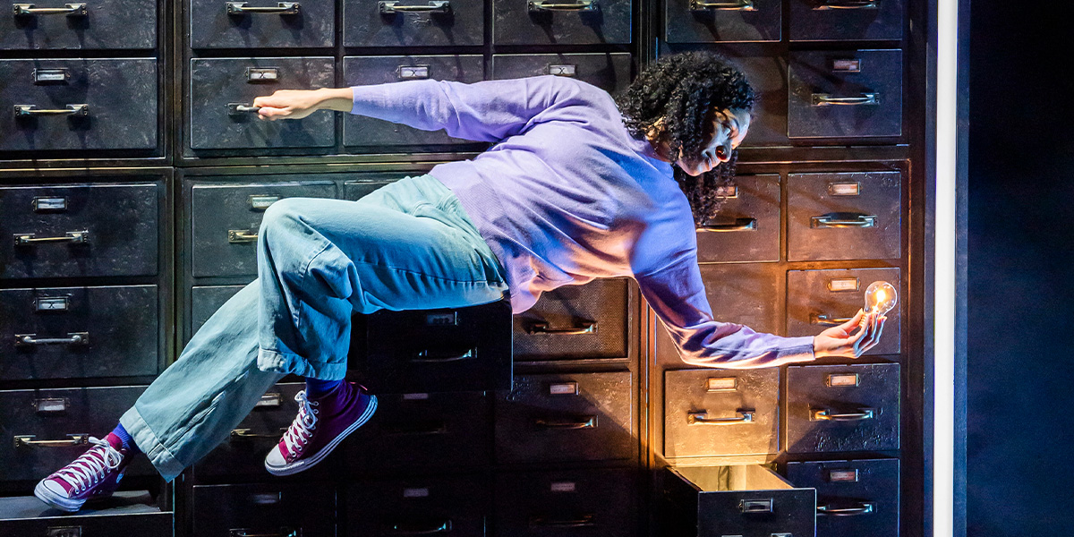 Four performers in vibrant costumes interact on stage, surrounded by dramatic lighting and abstract set designs.
