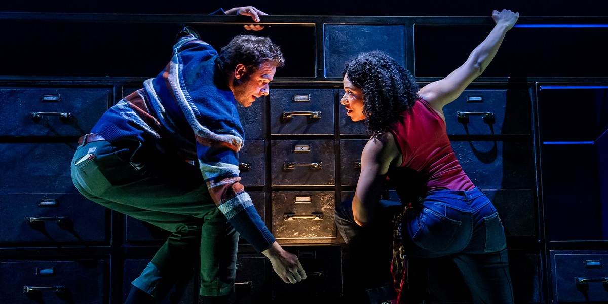 Four performers in vibrant costumes interact on stage, with dramatic lighting and a textured backdrop.