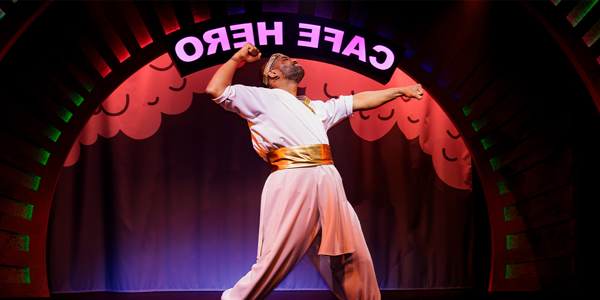 Stephan Boyce performs dramatically in a white costume with a gold belt, under a colourful arch.