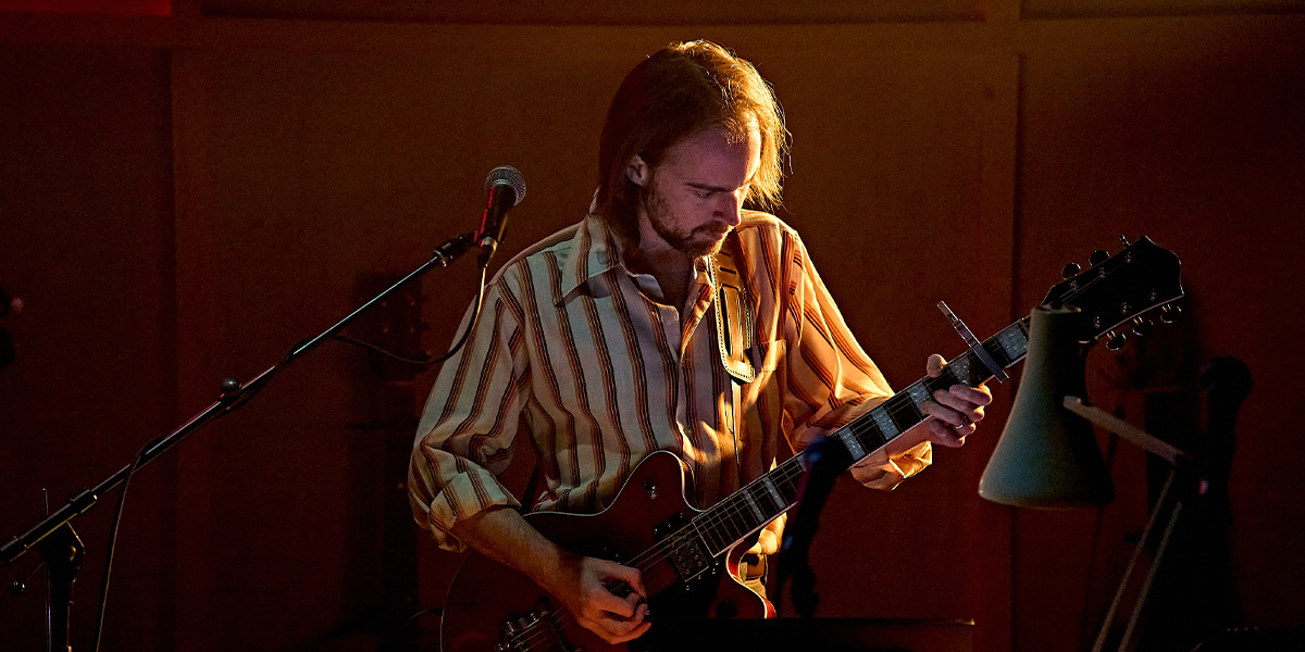 A man in a dark suit stands confidently, arms crossed, against a textured grey background.