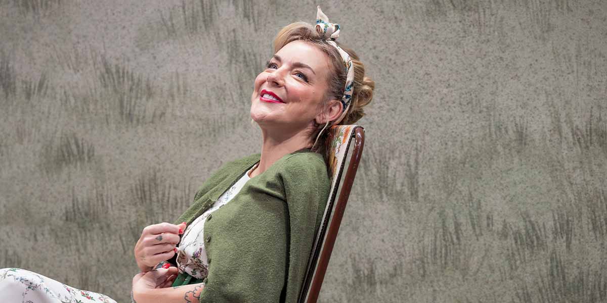 Sheridan Smith smiles while seated on a chair, wearing a floral dress and green cardigan.