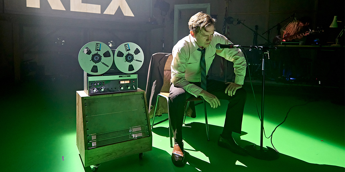 Jack Holden stands confidently in a dramatic pose, wearing a black outfit against a dark, textured background.