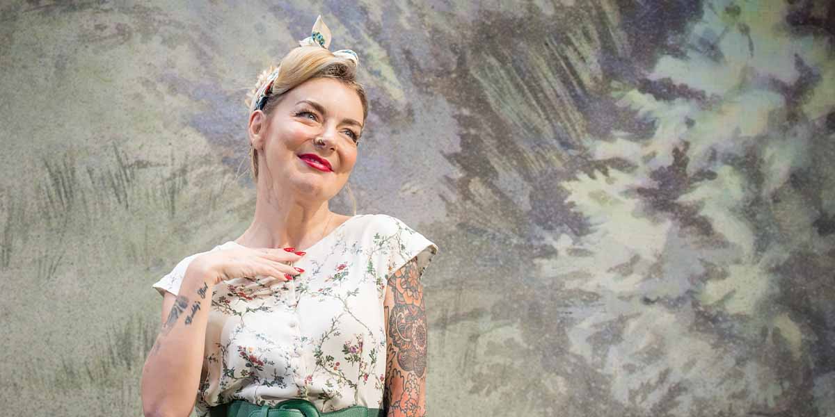 Sheridan Smith poses playfully, wearing a floral blouse and green belt, against a textured background.