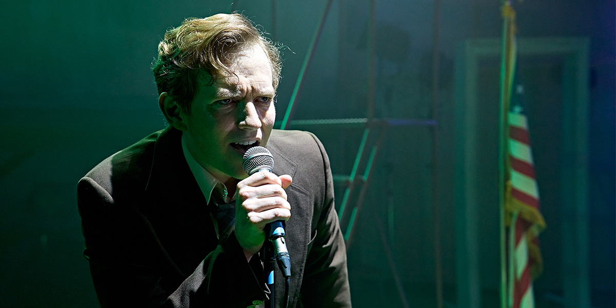 Jack Holden stands confidently in a dramatic pose, wearing a dark costume with textured details, against a muted background.