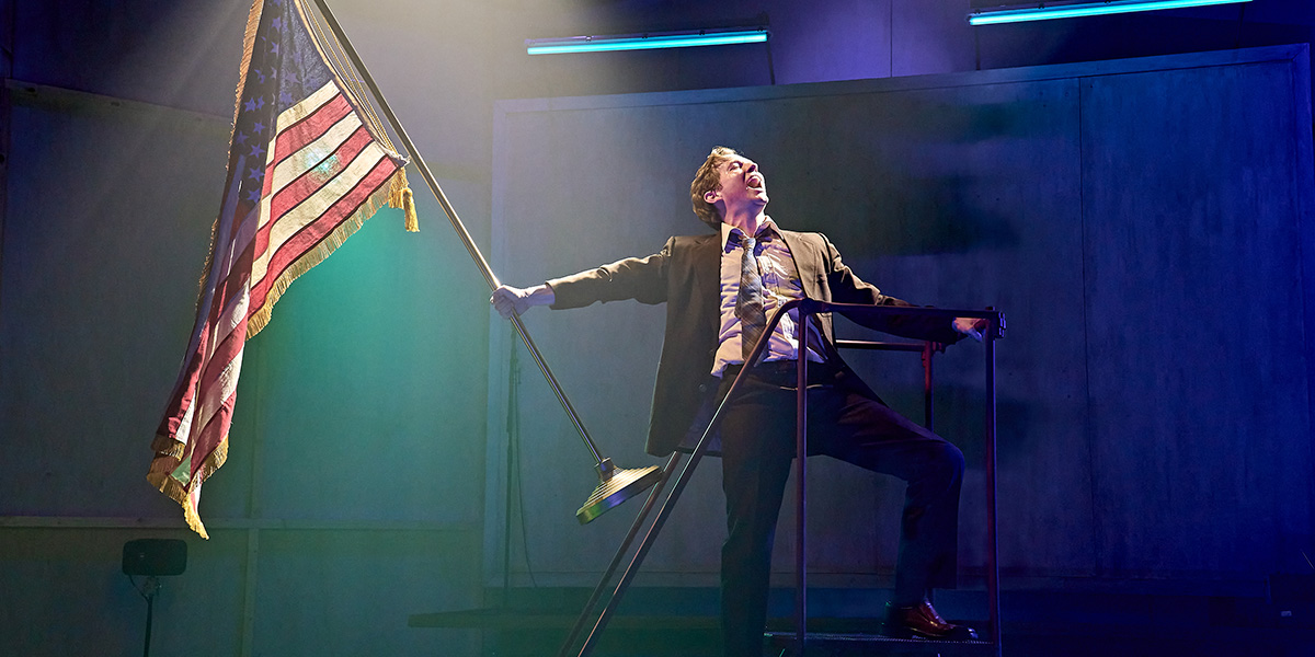 Jack Holden stands centre stage in a dramatic pose, wearing a dark costume against a dimly lit background.
