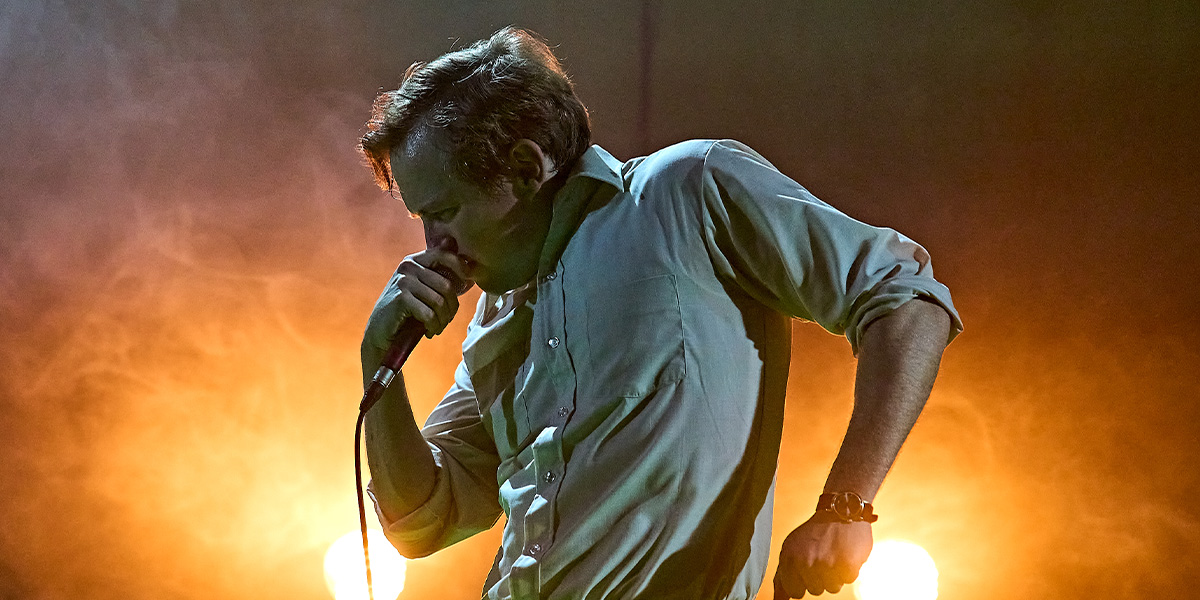 Jack Holden stands confidently in a dramatic pose, wearing a dark costume with textured details, against a muted background.
