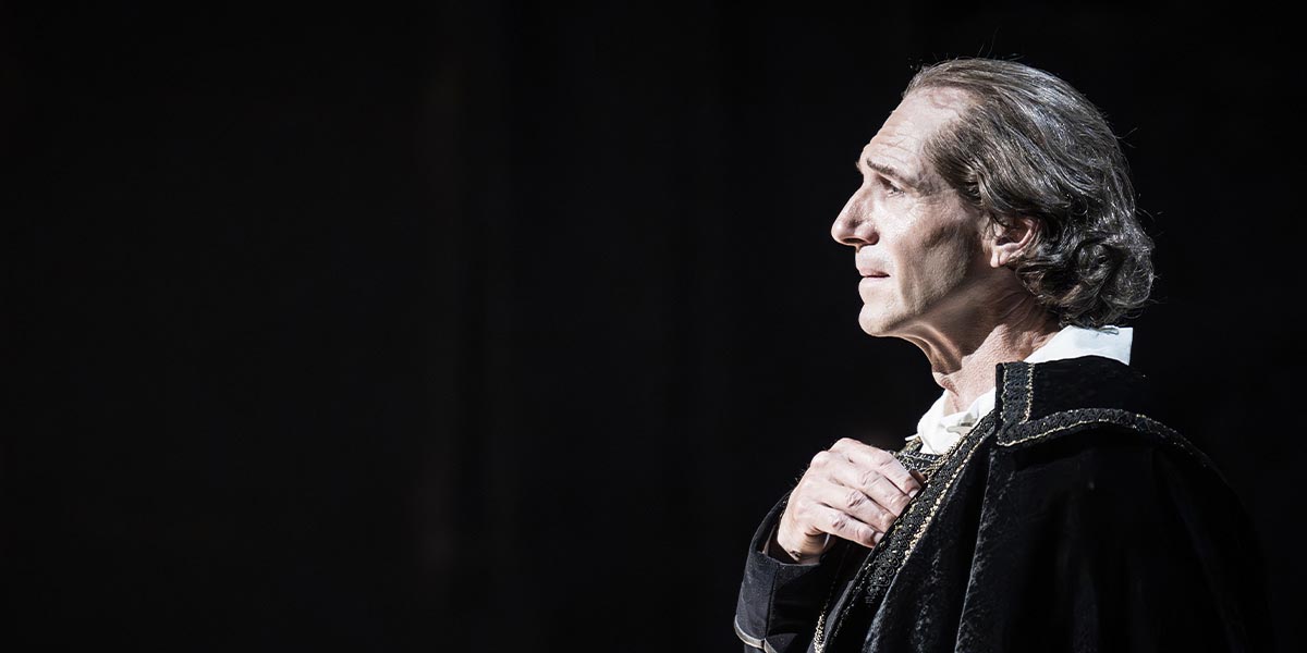 Ralph Fiennes, dressed in a dark costume, gazes thoughtfully while placing a hand on his chest.