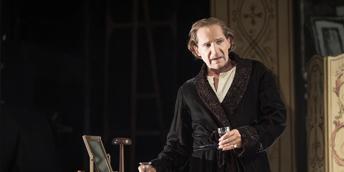 Ralph Fiennes, in a black robe, holds a glass while speaking, against a decorative background.