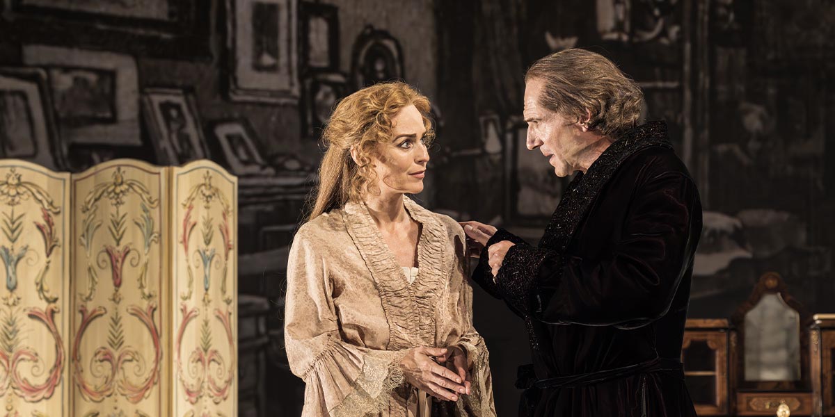 Ralph Fiennes and Miranda Raison pose together, smiling, against a neutral background with soft lighting.