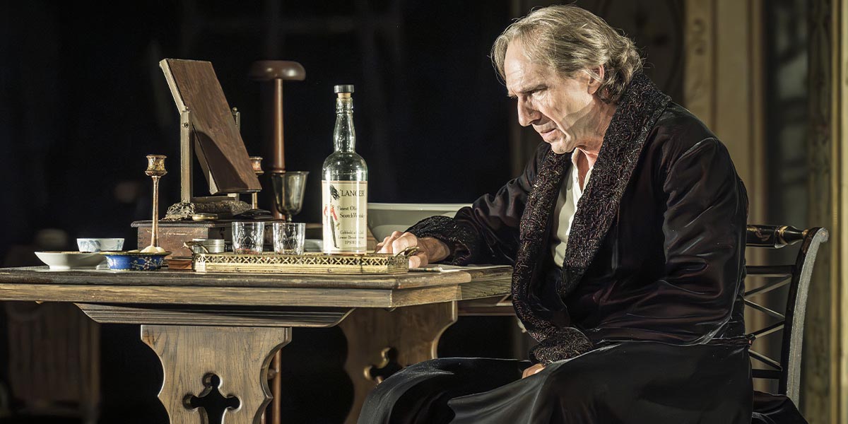 Ralph Fiennes portrays Henry Irving, dressed in period costume, with dramatic lighting and a dark background.