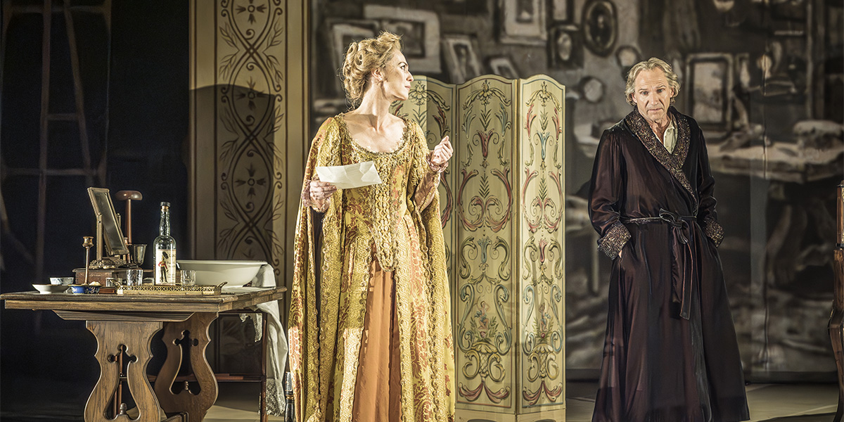 Ralph Fiennes and Miranda Raison sit together, engaged in conversation, with a blurred background of soft colours.