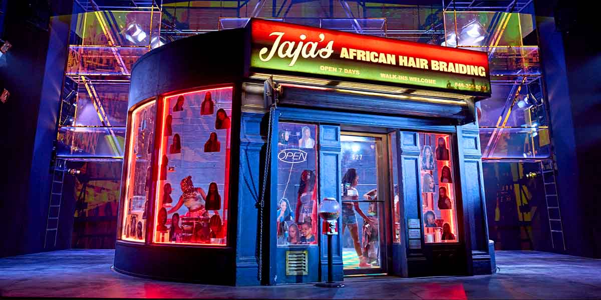 A stylist braids a client's hair in a salon, surrounded by hair products and vibrant decor.