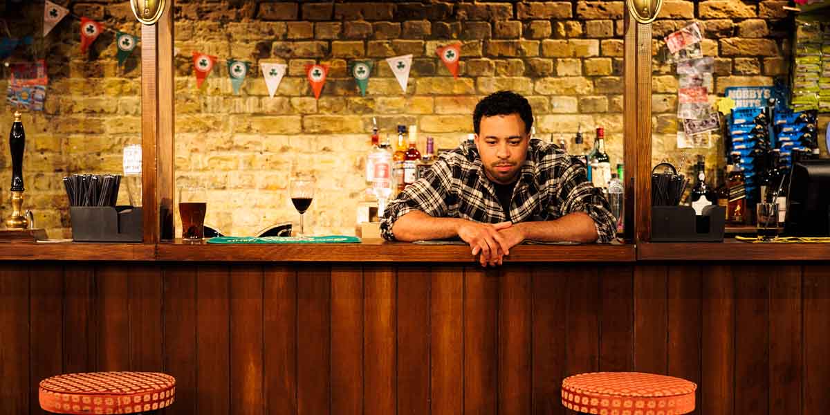 Aaron Anthony leans on a bar counter, wearing a checked shirt, with drinks and a brick wall behind.