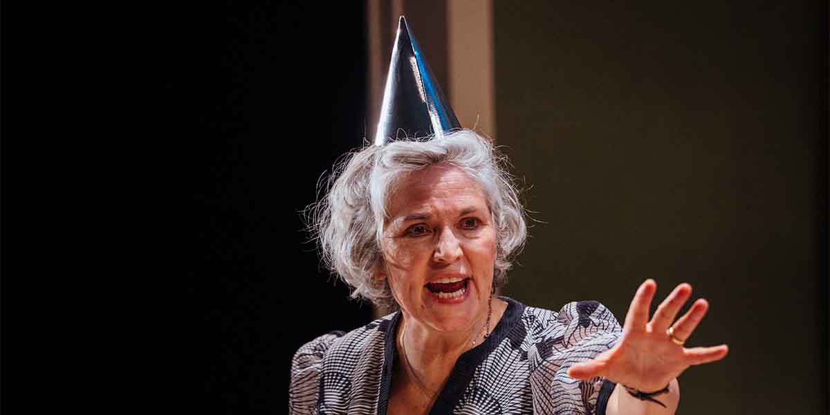 Andrea Irvine, wearing a party hat, gestures passionately while speaking against a dark background.