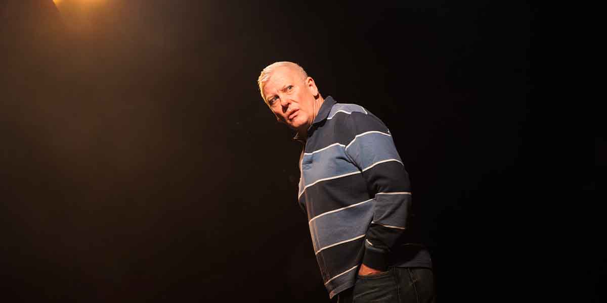 Deka Walmsley, wearing a striped blue shirt, stands in spotlight against a dark background, looking concerned.