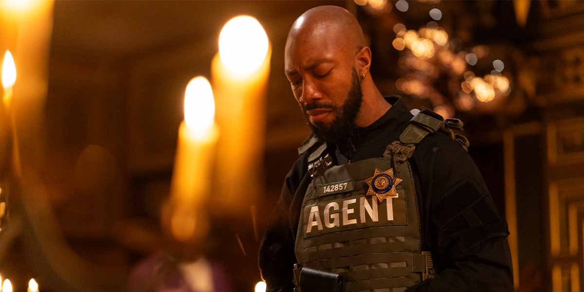 Elijah Cook as Tone, wearing a tactical vest labelled 'AGENT', stands solemnly amidst candlelight.