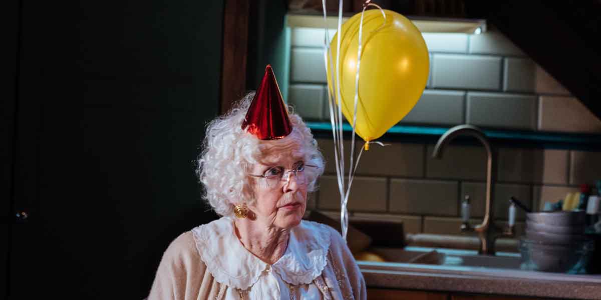 An elderly person with curly white hair wears a red party hat and holds a yellow balloon.
