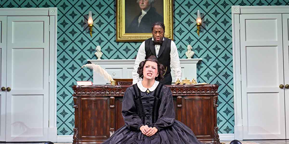 A woman in a black dress sits distressed at a desk, while a man in formal attire stands behind her.