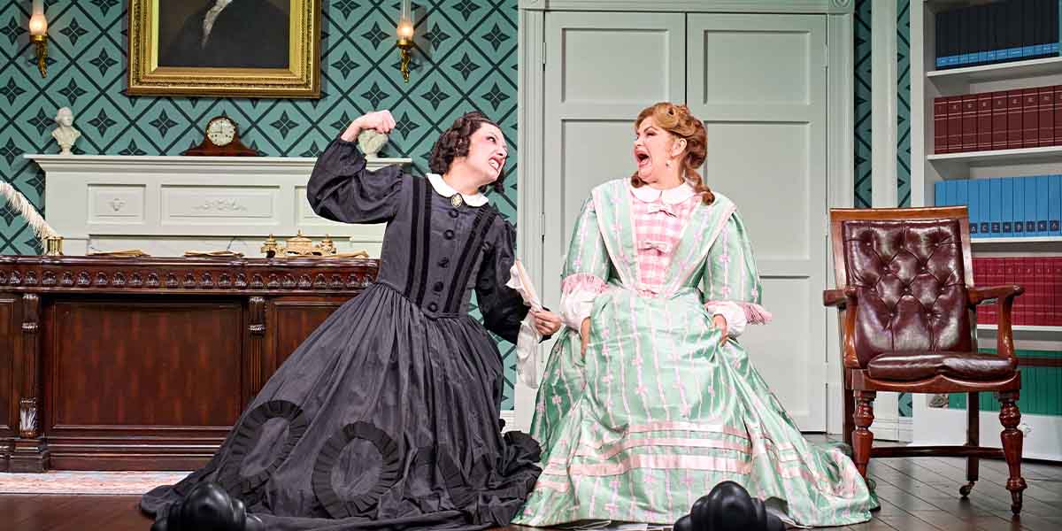 Two women in period costumes sit on the floor, laughing and gesturing playfully in a richly decorated room.